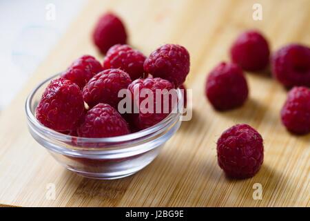Frisch und leicht unvollkommen Himbeeren in und um kleine Glasschale auf Holzbrett verschüttete Stockfoto