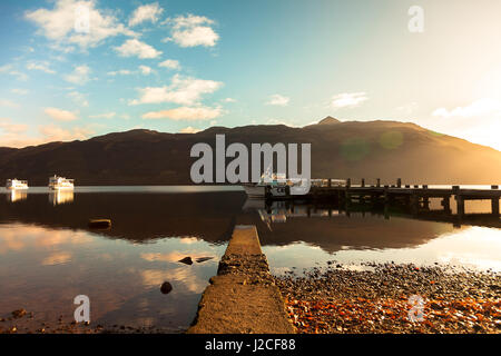 Sonnenuntergang auf dem noch Spiegel wie Wasser des Loch Lomond als Boot schwimmt am Ende eines Piers. Tarbet, Schottisches Hochland, Schottland Stockfoto