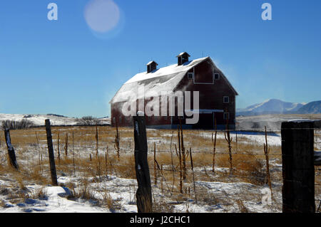 Schnee weht aus dem Dach einen rustikalen, hölzerne Scheune in Colorado.  Pole-Zaun läuft vor der Scheune.  Dünne Schicht Schnee sitzt auf Dach und Weide. Stockfoto