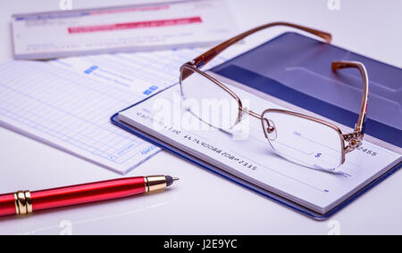 Bezahlung mit Scheck sofort, pünktlich. Gläser auf ein Scheckheft, roten Stift, finanzielle Dokumente im Hintergrund. closeup, Finanzkonzept. Stockfoto