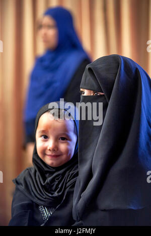 Eine muslimische Mutter trägt ein Gesicht bedeckende Burqua hält ihren kleinen Sohn in traditionelle orientalische Kleidung in einer Moschee in Anaheim, CA. Hinweis Frau im Hintergrund einen Hijab, traditionelle muslimische Frauen Kopftuch zu tragen. Stockfoto