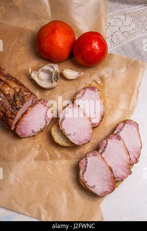 Scheiben mit Gewürzen und Kräutern Fleisch oder Schinken auf braunem Packpapier geräuchert. Tomaten, Knoblauch und kleinen Snacks von Brot und Fleisch und alte Vintage Messer. Braue Stockfoto