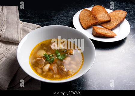 Bohnensuppe in Schüssel auf einem schwarzen Stein Hintergrund weiß. Einige toast auf weißen Teller und schwarze Mühle für Pfeffer auf braunem Tuch. Stockfoto