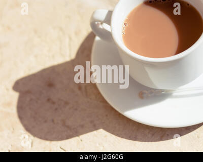 Tee mit Milch in eine weiße Tasse in der Morgensonne mit harten Schatten auf strukturierte Oberfläche des Holzes. Warme Töne. Stockfoto