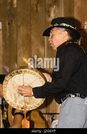 Athapaskische Mann singt Volkslieder und spielt eine Handtrommel während einer Versammlung in Alaska Native Heritage Center, Anchorage, AK Stockfoto