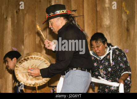 Athapaskische Mann singt Volkslieder und spielt eine Handtrommel mit seiner Familie während einer Versammlung in Alaska Native Heritage Center, Anchorage, AK Stockfoto