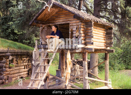 Athapaskische Frau sitzt auf der Plattform von einer traditionell Log gebaut Lebensmittel Lagerung Unterschlupf, genannt einen Cache befindet sich im Inneren ein Jagd- und Fischerei-Camp. Chena Dorf in Alaska Stockfoto