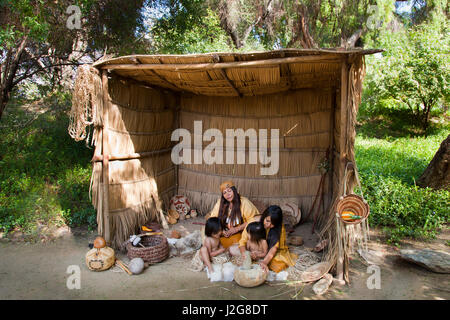 Acjachemen Nation Familie (Mission Indianer) Schleifen Eicheln unter einer möblierten Tule Reed Hütte für Schatten vor der Sonne am Nachmittag. Traditionellen Wohnhäuser der Indians of Southern California, befindet sich im Gegensatz Dorf Santa Fe Springs CA (MR) Stockfoto