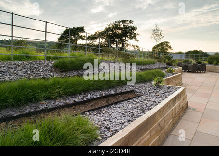 Schönen, gepflegten, privaten Garten mit modernem Design, gepflasterte Terrasse, Sträucher & Gräser in Linien auf erhöhte Grenze - Yorkshire, England, UK. Stockfoto