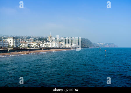 Blick auf Meer Teignmouth an einem sonnigen Frühlingstag mit Menschen zu Fuß entlang der Promenade. St. Michaels-Kirche steht im Vordergrund, im Hintergrund. Stockfoto