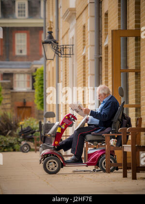 Chelsea Pensionär am Royal Hospital Chelsea saß auf Elektromobil außerhalb Wohnung lesen Zeitung, London, UK. Stockfoto