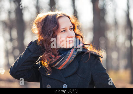 Lächelnde rothaarige Mädchen im Freien, weared Mantel und Schal. Vorfrühling im Park Ar Sonnenuntergang. Stockfoto