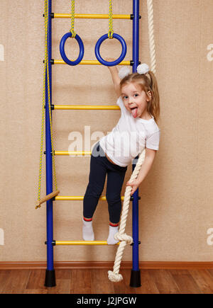 Kleines Mädchen in Sportbekleidung mit Zunge und steht auf der Leiter an der Sprossenwand. Das Konzept einer gesunden Lebensweise von einem jungen Alter. Kinder spor Stockfoto