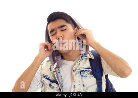 Hübscher junger Mann mit schwarzen Haaren in weißem T-shirt und Jean Jacke mit blauen Rucksack mit Kopfhörer und Gesang auf weißem Hintergrund im studio Stockfoto