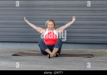 Ältere Frau, die Yoga-Übungen, wie sie auf einer Gymnastikmatte auf einem Holzdeck auf Gesundheit, Fitness und aktiven Lifestyle-Konzept sitzt zu passen Stockfoto