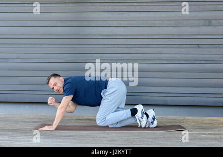 Erwachsener Mann stretching im Brett mit einem Bein und eine Hand heben und Blick in die Kamera, Seitenansicht vor grauem Hintergrund mit Textfreiraum Stockfoto