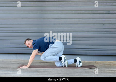 Erwachsener Mann stretching im Brett mit einem Bein und eine Hand heben und Blick in die Kamera, Seitenansicht vor grauem Hintergrund mit Textfreiraum Stockfoto