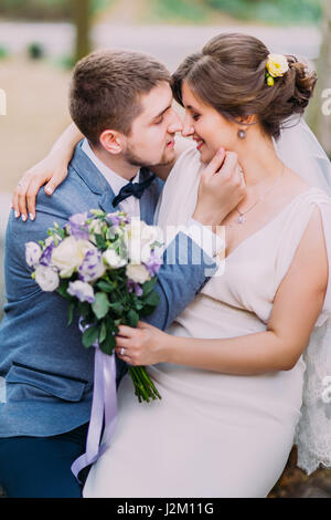 Elegante attraktive Brautpaar küssen im Freien. Berühren das Gesicht Braut, Bräutigam, während sie hält, Bridal rose Blumenstrauß in der hand Stockfoto