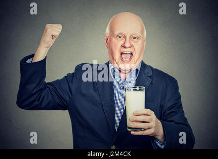 Heitere aufgeregt senior Gentleman hält ein Glas Milch und schaut in die Kamera auf grauem Hintergrund isoliert Stockfoto