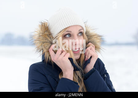 Glückliche junge Frau mit langen blonden Haaren tragen eine Fell getrimmt Jacke kuscheln in die Wärme des Kragens, wie steht sie auf der Seite in die Stockfoto