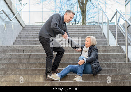 Mann hilft senior Frau aufzustehen, während sie auf Treppen im Freien in der Stadt sitzt, halten Sie ihr Knie Stockfoto