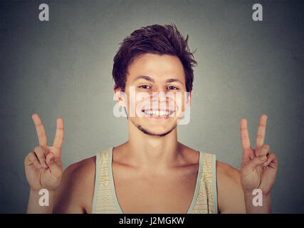 Junger Mann, gut aussehend Student hält Frieden Sieg, zwei Zeichen isoliert grau hinterlegt. Positive menschliche Emotion Gesichtsausdruck Symbole Haltung. Li Stockfoto