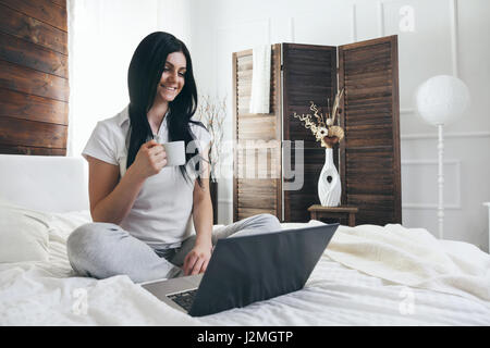 Entspannen auf dem Bett. Schöne junge Frau, eine Tasse Kaffee zu genießen und mit ihrem laptop Stockfoto