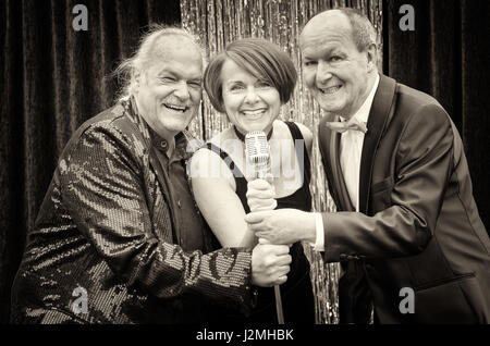 Drei glückliche Sänger Musizieren auf der Bühne mit einer temperamentvollen Frau, flankiert von zwei ältere Männer lachen und Lächeln vor dem Mikrofon in Stockfoto