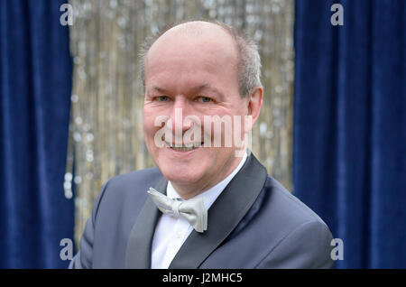 Attraktive senior Glatze Mann in eine Fliege und Jacke posiert vor einem blauen Vorhang mit Silberflitter mit einem freundlichen Lächeln in die Kamera schaut Stockfoto