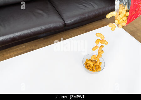 Erdnuß Locken fallen aus einer Packung Chips in eine Glasschüssel auf weißen Couchtisch Stockfoto
