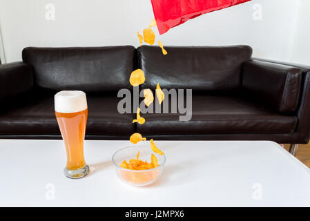 Kartoffel-Chips fallen aus einer Packung Chips in eine Glasschüssel mit Weißbier auf weißen couch Tisch Stockfoto