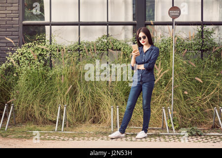 junge asiatische Frau, trägt blaue Jeans und Sonnenbrille, draußen mit ihrem Smartphone, Vintage Farbe Stil Stockfoto