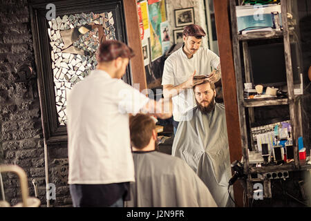 bärtiger Mann immer Haarschnitt von Friseur beim Sitzen im Stuhl bei barbershop Stockfoto