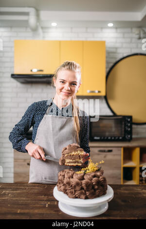 Frau Koch hält Stück Schokoladenkuchen auf der Klinge, kulinarisches Meisterwerk. Küche im Hintergrund. Hausgemachte Süßspeise Stockfoto