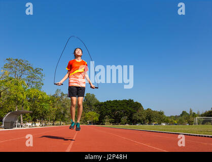 junge asiatische Sportlerin Seilspringen im Freien auf der Laufstrecke am Fußball oder "Fußball", Feld mit Bäumen und klaren blauen Himmel im Hintergrund Stockfoto