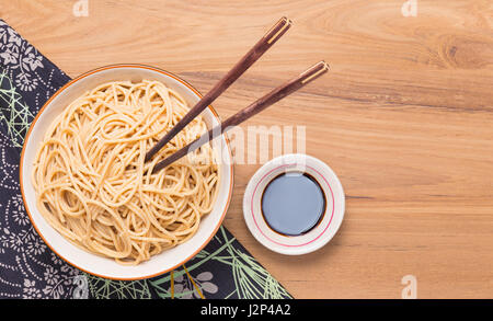 Schüssel Nudeln mit hölzernen Stäbchen und eine Tasse Sojasauce auf japanische Art Tischdecke auf Holztisch, Raum für Exemplar Stockfoto