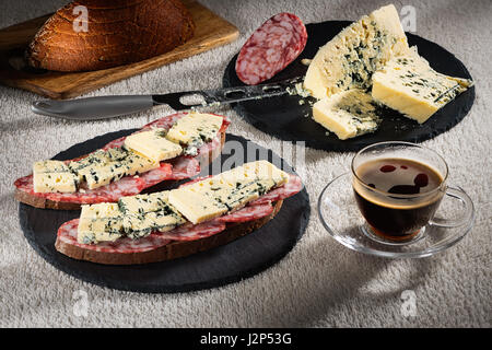 Sandwiches mit Käse Dorblu und geräucherte Rohwurst auf schwarzem Schiefer Platten, ein Käsemesser und eine Tasse heißen Kaffee Stockfoto