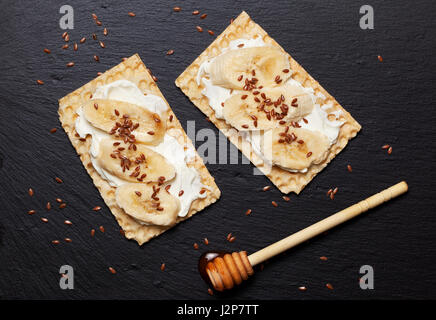 Ernährung, gesundes Frühstück. Knuspriges Brot Sandwich, Ricotta, Banane, Honig und Leinsamen auf schwarzem Schiefer Hintergrund Stockfoto