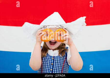 Niederländische Mädchen tragen traditionelle Tracht, Kleid und Hut hält orange Donuts auf der Flagge der Niederlande. Kind mit Oranje Souvenirs fr Stockfoto