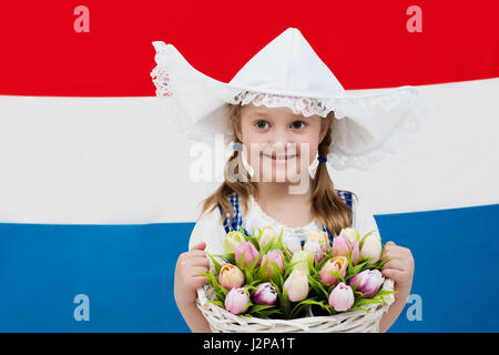 Niederländische Mädchen tragen traditionelle Tracht, Kleid und Hut hält Korb mit Tulpen auf der Flagge der Niederlande. Kind mit Souvenirs aus H Stockfoto