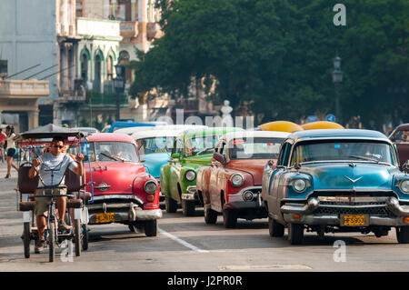 Havanna - Juni 2011: Eine einsame Rikscha, bekannt als ein Bicitaxi teilt sich die Straße mit einer langen Reihe von bunten klassischen amerikanischen Autos aus den 1950er Jahren. Stockfoto