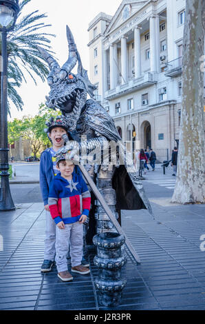 Ein paar der jungen Brüder Pose für ein Foto mit einem Straßenkünstler gekleidet wie eine silberne Drache auf La Rambla in Barcelona, Spanien. Stockfoto