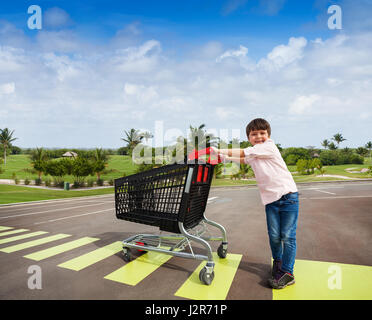 Porträt von Kid Boy beim Überqueren der Straße am Zebrastreifen der Supermarkt-Parkplatz mit Warenkorb Stockfoto