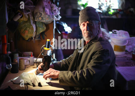 Ein senior erwachsenen Mann in die Kamera schaut und ein Stück Stoff auf eine Nähmaschine nähen. Stockfoto