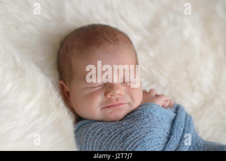 Schlafen, neun Tage alten, neugeborenes Baby Boy mit einem Lächeln auf seinem Gesicht. Er ist in einer leichten blauen Packung gepuckt. Gedreht im Studio auf einem weißen, Schaffell-Teppich. Stockfoto