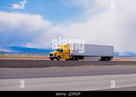 Klassische moderne Semi Truck leuchtend gelbe Farbe mit einer weißen voller Länge Anhänger aerodynamische Röcke auf einer flachen Autobahn in Utah mit den Blue Mountains auf Stockfoto