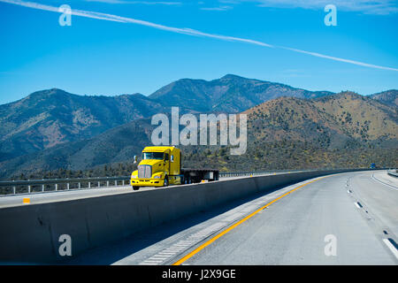 Hellgelb, leistungsstarke Sattelzug Sattelschlepper mit einem Tieflader auf dem interstate Berg pass Autobahn wiederum mit den Berggipfeln und blauer Himmel. Stockfoto