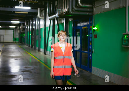 weibliche Angestellte in einer orangefarbenen Robe-Weste im Arbeitsraum einer Produktionsstätte, übt die Kontrolle im Lager Produktionsflächen Stockfoto