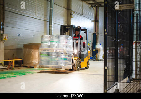 Lagermitarbeiter im Overall Kleidung, Fahrer Ausstellungsgeräte fleißig an Logistik Moving on Loader Paletten mit Bierfässer auf Linie der Fabrik für Pro Stockfoto