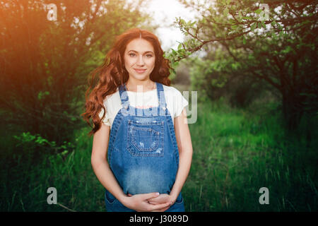 Junge schöne schwangere Mädchen mit langen braunen Haaren in insgesamt Jeans im grünen Garten. Stockfoto